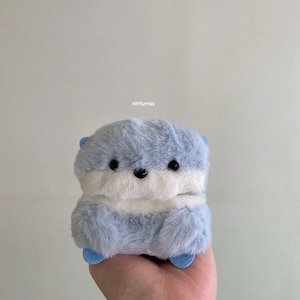 May include: A plush toy shaped like an otter with light blue and white faux fur. The creature has small black eyes, a black nose, and two small blue felt ears. The text "ciitfurries" is printed in small white letters above the plush toy.