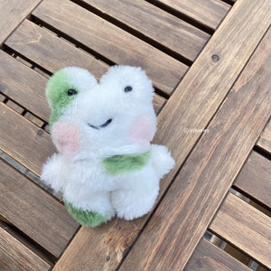 以下が含まれることがあります： 緑とピンクのアクセントが付いた白いぬいぐるみのかえる。かえるは幸せそうな表情をしていて、木製の表面に座っています。