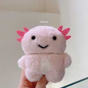 May include: A plush pink axolotl-shaped case with black eyes and a smiling mouth. The case has pink frills on the head and small arms. The text "clitfurries" is visible in the background.