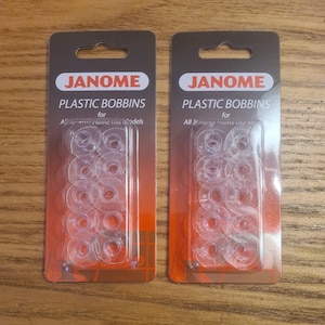 May include: Two packs of clear plastic bobbins for Janome sewing machines. Each pack contains 12 bobbins and has a red label with the text "JANOME PLASTIC BOBBINS for All Janome Home Use Models".