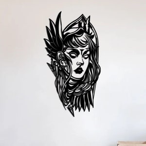May include: Black metal wall art depicting a woman's face with an elaborate headdress and feathered wings. The artwork is mounted on a white wall in a modern living room setting. The woman's face has a detailed design with closed eyes and dark lips.