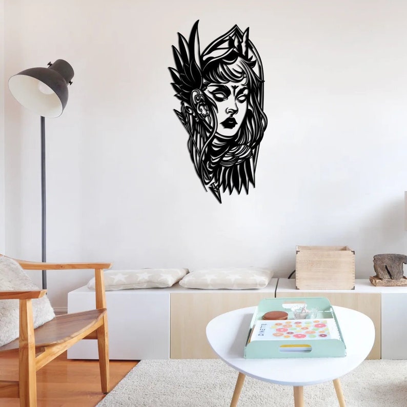 May include: Black metal wall art depicting a woman's face with an elaborate headdress and feathered wings. The artwork is mounted on a white wall in a modern living room setting. The woman's face has a detailed design with closed eyes and dark lips.