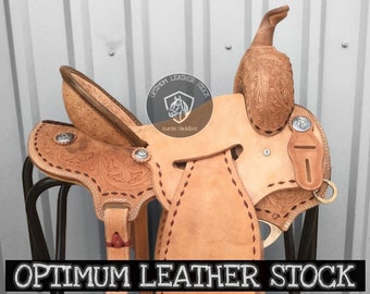 May include: A tan leather saddle with intricate tooling and stitching. The saddle is on a black metal stand. The saddle is labeled "Optimum Leather Stock Horse Saddles".