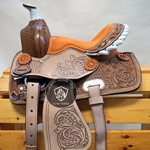 May include: A brown leather horse saddle with intricate floral tooling. The saddle features a white horn, orange suede seat, and silver accents. The saddle is displayed on a wooden stand.