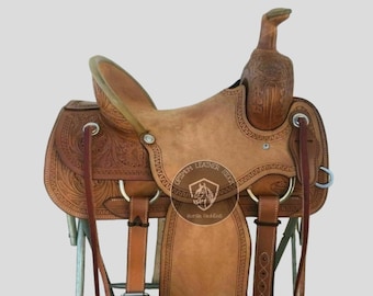 May include: A brown leather saddle with intricate tooling and stitching. The saddle features a high cantle and a  horn with a decorative design. The saddle is on a metal stand.