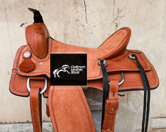 May include: A brown leather saddle with a tooled design and black stitching. The saddle has a saddle horn, stirrups, and a cinch. The saddle is sitting on a blue stool. The saddle is labeled "Optimum Leather Stock".