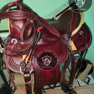 K&ouml;nnte beinhalten: Ein brauner Ledersattel f&uuml;r Pferde mit aufwendigen N&auml;hten und einem gelben Lederbesatz. Der Sattel ist mit einem silbernen Koncho verziert und hat einen Ledersteigb&uuml;gel. Der Sattel ist mit "Optimum Leather Stock Horse Saddles" beschriftet.