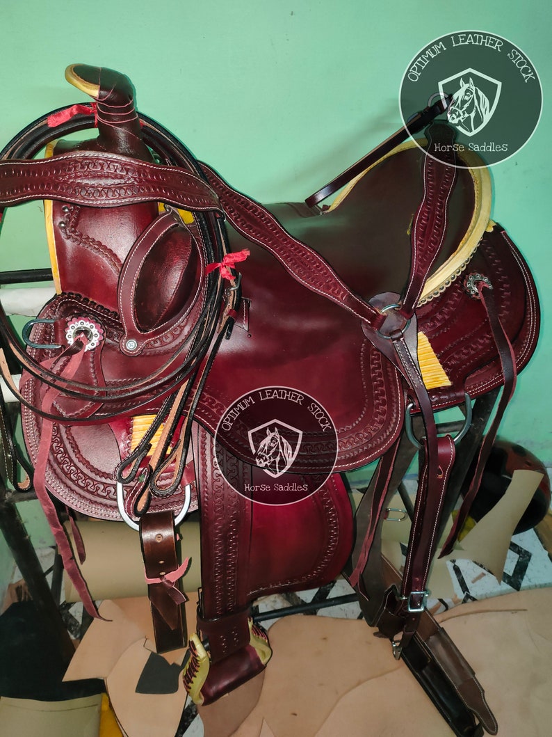 K&ouml;nnte beinhalten: Ein brauner Ledersattel f&uuml;r Pferde mit aufwendigen N&auml;hten und einem gelben Lederbesatz. Der Sattel ist mit einem silbernen Koncho verziert und hat einen Ledersteigb&uuml;gel. Der Sattel ist mit "Optimum Leather Stock Horse Saddles" beschriftet.