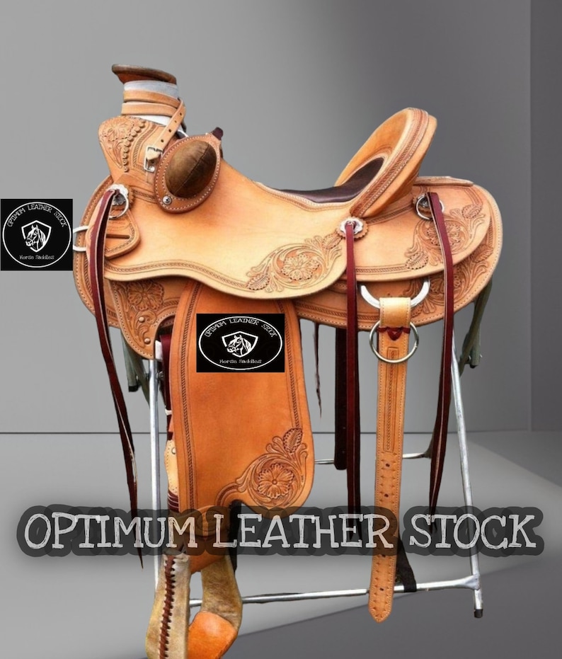 May include: A brown leather saddle with intricate tooling and a saddle horn. The saddle is on a metal stand and has a "OPTIMUM LEATHER SEAT" brand on the flap.
