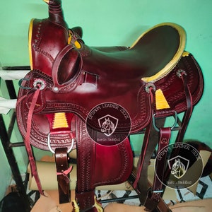 K&ouml;nnte beinhalten: Ein brauner Ledersattel f&uuml;r Pferde mit gelben N&auml;hten und einem Sattelhorn. Der Sattel ist mit einem gepr&auml;gten Lederdesign verziert und hat einen Metallsteigb&uuml;gel. Der Sattel ist mit "Optimum Leather Stock Horse Saddles" beschriftet.