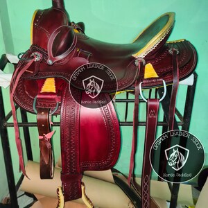 K&ouml;nnte beinhalten: Ein burgunderfarbener Ledersattel mit gelben N&auml;hten und einem goldfarbenen Horn. Der Sattel steht auf einem Holzst&auml;nder und hat einen Lederriemen mit einer Metallschnalle. Der Sattel ist mit "Optimum Leather Stock Horse Saddles" beschriftet.