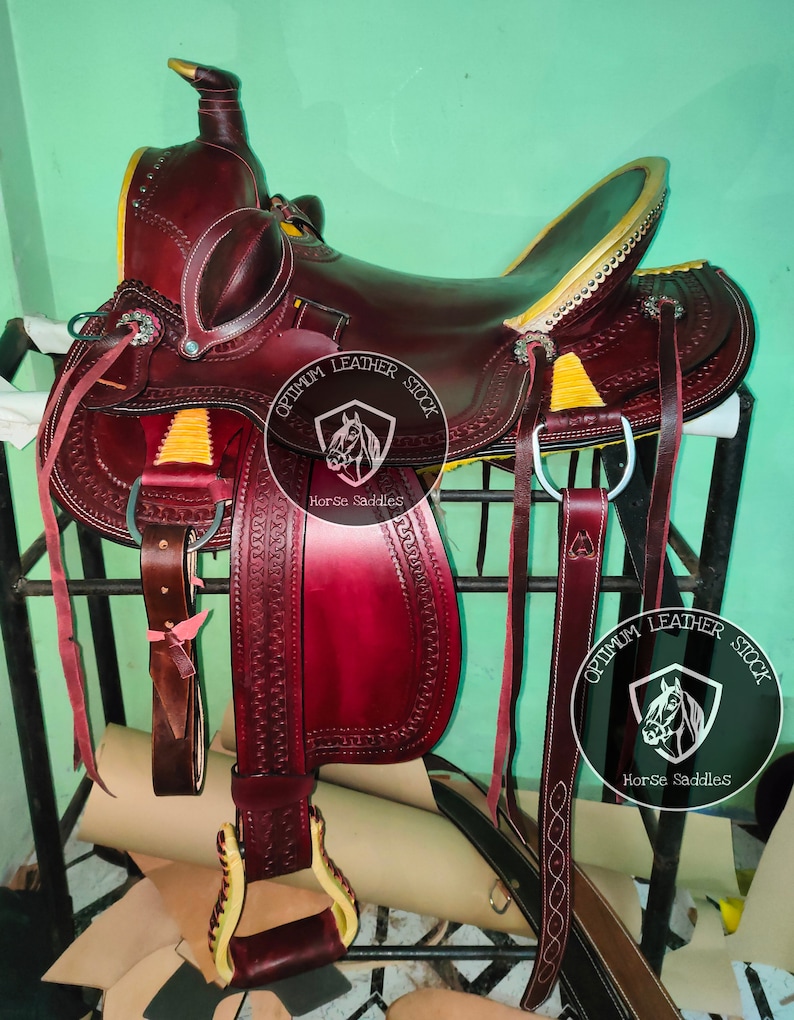 K&ouml;nnte beinhalten: Ein burgunderfarbener Ledersattel mit gelben N&auml;hten und einem goldfarbenen Horn. Der Sattel steht auf einem Holzst&auml;nder und hat einen Lederriemen mit einer Metallschnalle. Der Sattel ist mit "Optimum Leather Stock Horse Saddles" beschriftet.