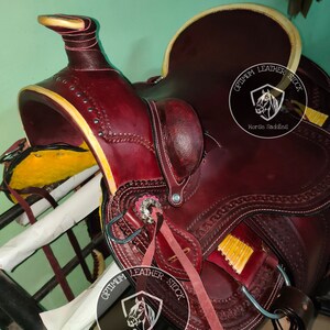 K&ouml;nnte beinhalten: Ein burgunderfarbener Ledersattel mit aufwendigen N&auml;hten und gelbem Besatz. Der Sattel verf&uuml;gt &uuml;ber einen silbernen Concho und einen Lederriemen mit dem Text "Optimum Leather Stock Horse Saddles".