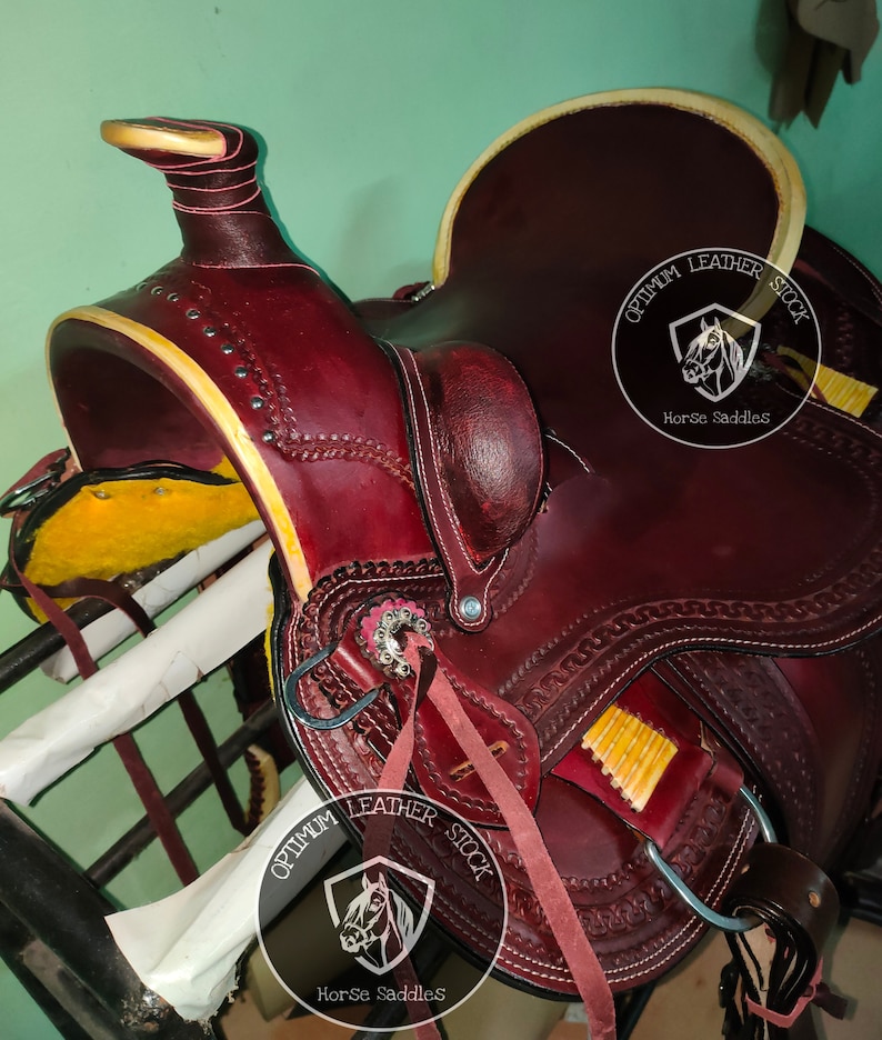 K&ouml;nnte beinhalten: Ein burgunderfarbener Ledersattel mit aufwendigen N&auml;hten und gelbem Besatz. Der Sattel verf&uuml;gt &uuml;ber einen silbernen Concho und einen Lederriemen mit dem Text "Optimum Leather Stock Horse Saddles".