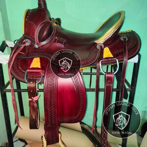 K&ouml;nnte beinhalten: Ein burgunderfarbener Ledersattel mit goldenen Akzenten und N&auml;hten. Der Sattel steht auf einem Metallst&auml;nder und hat einen Ledersteigb&uuml;gel. Der Sattel ist mit "Optimum Leather Stock Horse Saddles" beschriftet.