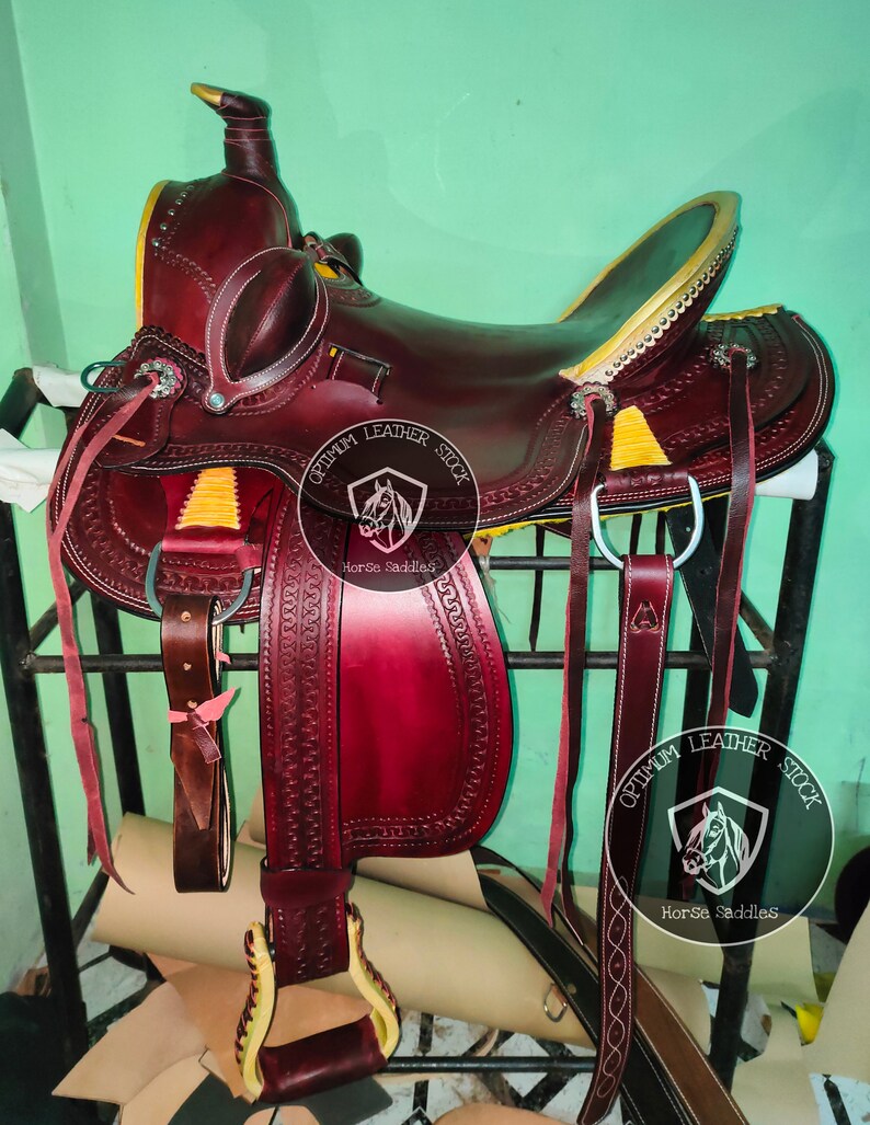 K&ouml;nnte beinhalten: Ein burgunderfarbener Ledersattel mit goldenen Akzenten und N&auml;hten. Der Sattel steht auf einem Metallst&auml;nder und hat einen Ledersteigb&uuml;gel. Der Sattel ist mit "Optimum Leather Stock Horse Saddles" beschriftet.