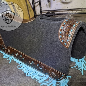 May include: A grey saddle pad with brown leather trim and turquoise fringe. The leather trim features a floral design. The saddle pad is labelled "Optimum Leather Stock Horse Saddles".