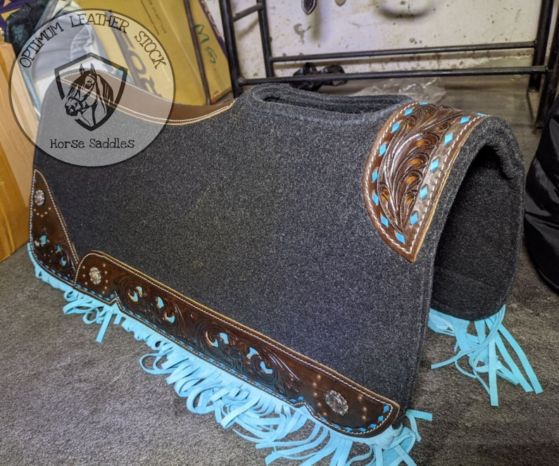 May include: A grey saddle pad with brown leather trim and turquoise fringe. The leather trim features a floral design. The saddle pad is labelled "Optimum Leather Stock Horse Saddles".