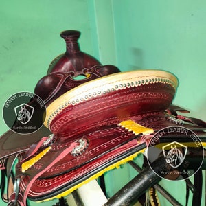 K&ouml;nnte beinhalten: Nahaufnahme eines burgunderfarbenen Ledersattels mit gelben N&auml;hten und silbernen Akzenten. Der Sattel ist mit aufwendigen Verzierungen verziert und tr&auml;gt das Logo "Optimum Leather Stock Horse Saddles" an der Seite.