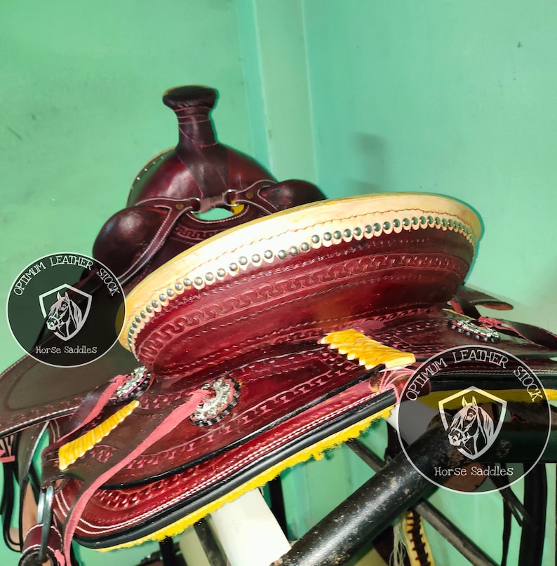 K&ouml;nnte beinhalten: Nahaufnahme eines burgunderfarbenen Ledersattels mit gelben N&auml;hten und silbernen Akzenten. Der Sattel ist mit aufwendigen Verzierungen verziert und tr&auml;gt das Logo "Optimum Leather Stock Horse Saddles" an der Seite.