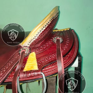 K&ouml;nnte beinhalten: Nahaufnahme eines roten Ledersattels mit gelben Akzenten und einem silbernen Steigb&uuml;gel. Der Sattel ist mit aufwendigen N&auml;hten verziert und tr&auml;gt das Logo "Optimum Leather Stock Horse Saddles" an der Seite.