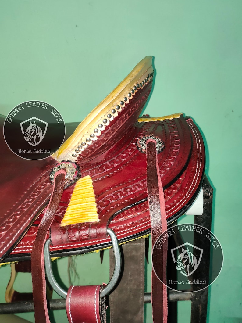 K&ouml;nnte beinhalten: Nahaufnahme eines roten Ledersattels mit gelben Akzenten und einem silbernen Steigb&uuml;gel. Der Sattel ist mit aufwendigen N&auml;hten verziert und tr&auml;gt das Logo "Optimum Leather Stock Horse Saddles" an der Seite.
