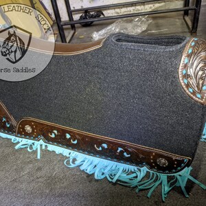 May include: A grey saddle pad with brown leather trim and turquoise fringe. The pad features a decorative floral design on the leather trim. The pad is labelled "Optimum Leather Stock Horse Saddles".