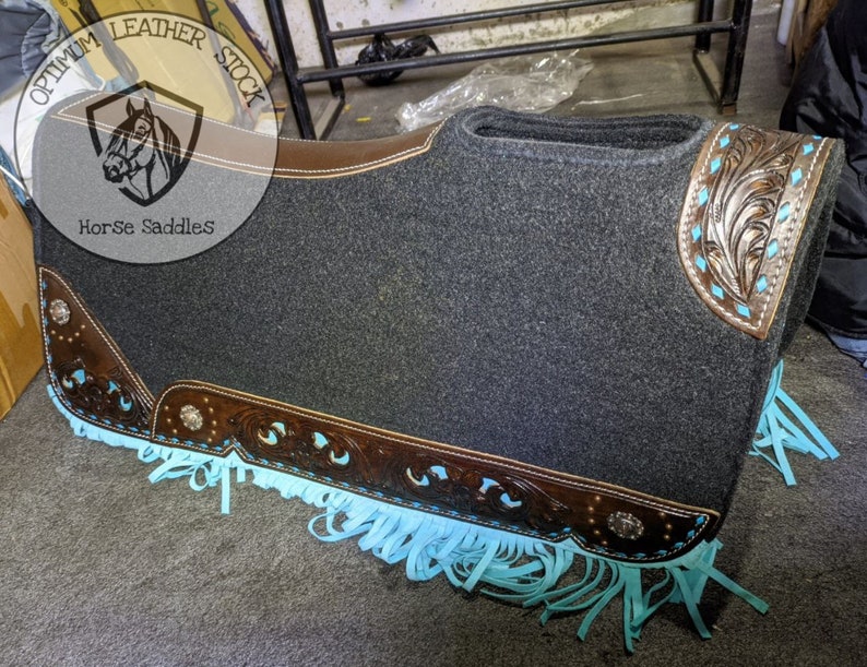 May include: A grey saddle pad with brown leather trim and turquoise fringe. The pad features a decorative floral design on the leather trim. The pad is labelled "Optimum Leather Stock Horse Saddles".