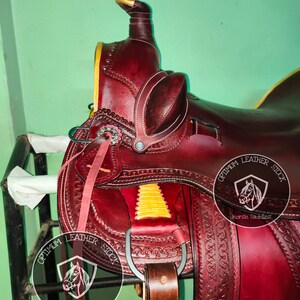 K&ouml;nnte beinhalten: Ein burgunderfarbener Ledersattel mit aufwendigen N&auml;hten und einem gelben Akzent. Der Sattel ist mit zwei Logos verziert, auf denen "Optimum Leather Stock Horse Saddles" steht.