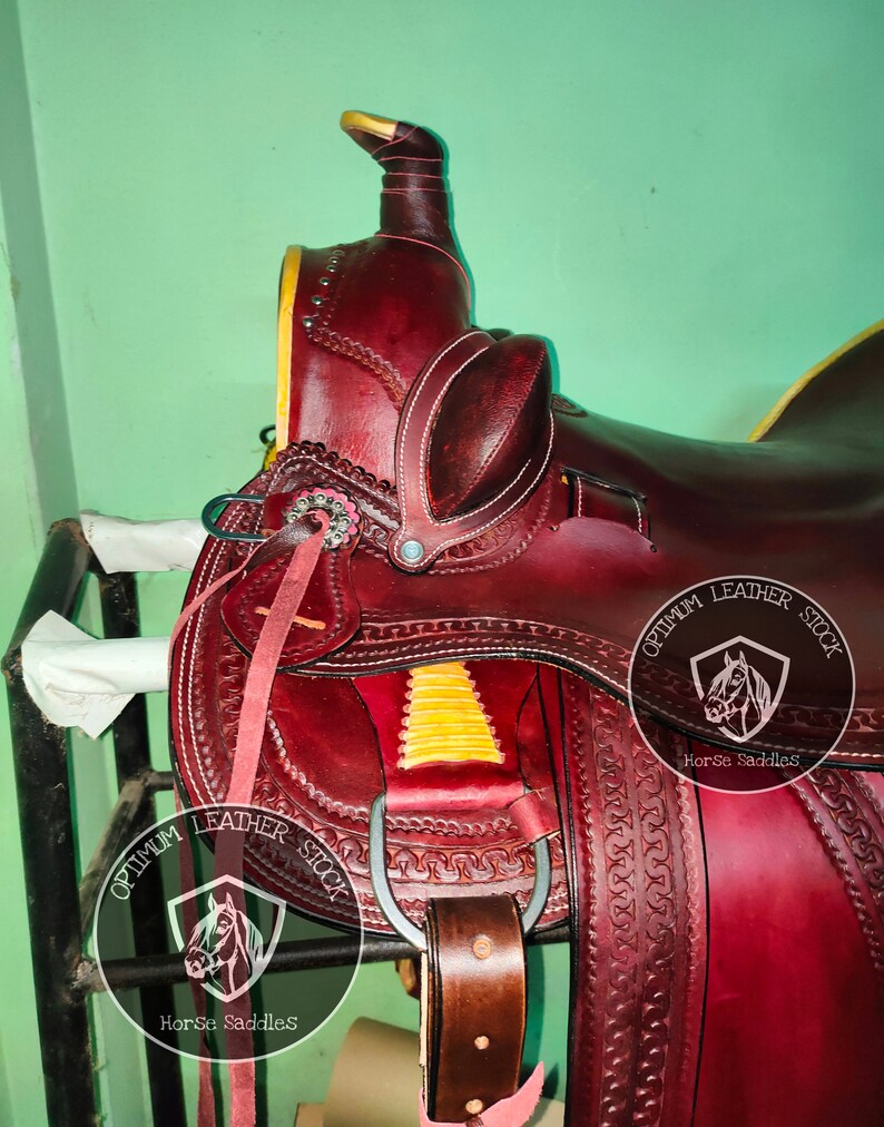 K&ouml;nnte beinhalten: Ein burgunderfarbener Ledersattel mit aufwendigen N&auml;hten und einem gelben Akzent. Der Sattel ist mit zwei Logos verziert, auf denen "Optimum Leather Stock Horse Saddles" steht.