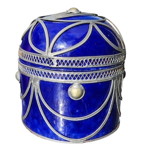 May include: A cylindrical cobalt blue container with a domed lid, featuring intricate silver-toned metal filigree overlays in a repeating petal and swag pattern, accented with small circular metal studs along the midsection and top.