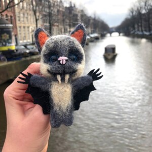 May include: A hand-crafted felt bat with black wings, a white furry body, and orange ears. The bat has a toothy grin and blue eyes. The bat is held in front of a blurry background of a canal.