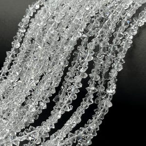 Peut inclure: Plusieurs brins de perles en cristal transparent, facettées et en forme de diamant, disposées en lignes parallèles. Chaque perle présente une taille géométrique nette qui reflète la lumière, créant un aspect brillant et étincelant sur l'ensemble.