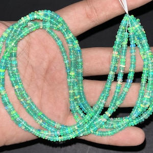 May include: A strand of green opal beads, with a slight iridescent shimmer, is displayed on a hand. The beads are small and round, and the strand is long enough to be worn as a necklace.
