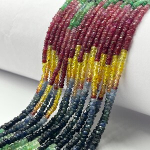 Puede incluir: Múltiples hilos de cuentas de piedras preciosas facetadas dispuestas en un patrón degradado. Las cuentas transicionan a través de colores que incluyen verde vibrante, rojo intenso, amarillo brillante, naranja suave, azul claro, azul marino y negro.