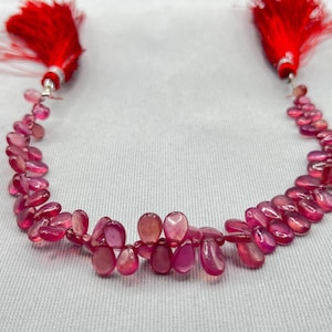 May include: A necklace composed of pink, teardrop-shaped gemstones. The gemstones are strung together, creating a curved design. Red tassels are attached at each end of the necklace. The necklace is presented on a white surface.