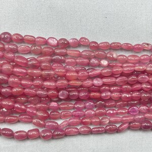 May include: Strands of pink gemstone beads. The beads are oval-shaped and have a polished, glossy finish. The color varies from light to deep pink, with some beads appearing translucent. The beads are strung together, ready for jewelry making.