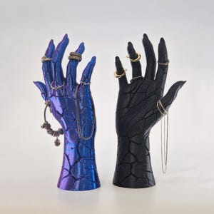 May include: Two hand-shaped jewelry holders, one blue and one black, with a cracked texture. Both hands are adorned with rings and necklaces.