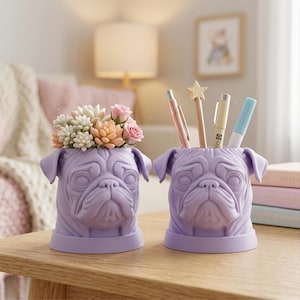 Puede incluir: Dos organizadores de escritorio con forma de carlino lavanda. Uno contiene bolígrafos y lápices, el otro tiene suculentas y rosas artificiales. Los organizadores están sobre una mesa de madera, con un fondo suave y borroso.