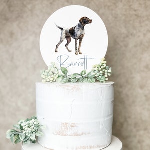 May include: A two-tiered white cake with a round topper featuring a watercolor illustration of a brown and white dog with the name "Barrett" written in cursive.