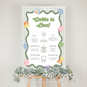 May include: A personalized birthday milestone poster with a green and white gingham border. The poster features the text "Callie is One!" and includes sections for favorite things, height, weight, nicknames, and favorite foods. The poster is displayed on a wooden easel.