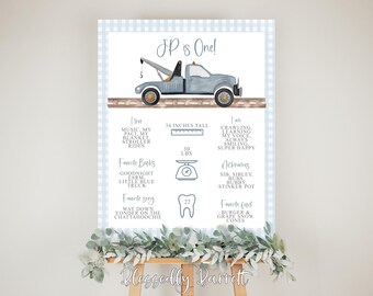 May include: A blue and white checkered poster with a watercolor illustration of a blue tow truck. The poster is a first birthday milestone sign for a baby boy named JP. It includes details like his height, weight, favorite books, songs, and nicknames.