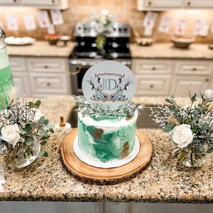 K&ouml;nnte beinhalten: Ein gr&uuml;ner und goldener Kuchen mit einem wei&szlig;en Topper, auf dem "B.D." steht und der V&ouml;gel und Gr&uuml;npflanzen zeigt. Der Kuchen steht auf einem Holzkuchenst&auml;nder und ist von Gr&uuml;npflanzen und wei&szlig;en Blumen umgeben.
