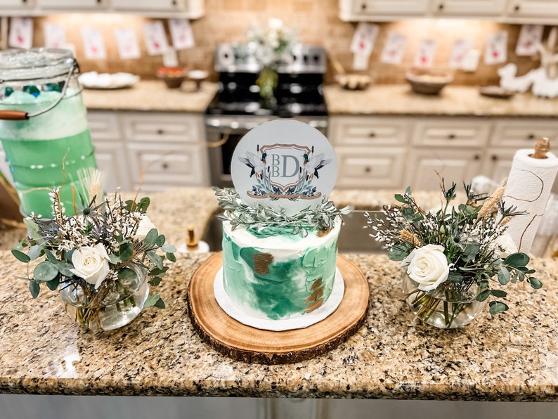 K&ouml;nnte beinhalten: Ein gr&uuml;ner und goldener Kuchen mit einem wei&szlig;en Topper, auf dem "B.D." steht und der V&ouml;gel und Gr&uuml;npflanzen zeigt. Der Kuchen steht auf einem Holzkuchenst&auml;nder und ist von Gr&uuml;npflanzen und wei&szlig;en Blumen umgeben.