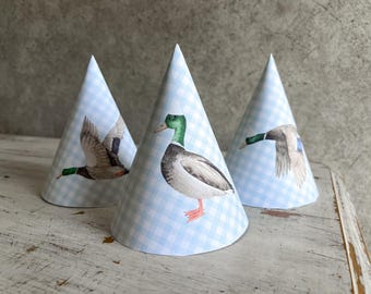May include: Three blue and white party hats with a gingham pattern. Each hat features a different illustration of a duck. One duck is flying, one is swimming, and one is standing on the water.