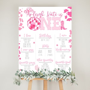 May include: A pink and white printable birthday sign for a one-year-old girl. The sign features a disco ball, cowboy boots, and a pink and white cow print pattern. The sign includes the girl's name, Arleigh Kate, and her birthday, July 14, 2022. It also lists her favorite things, including mommy kisses, bath time, and her favorite songs.