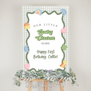 May include: A birthday sign with the text "OUR LITTLE Lucky Charm IS ONE Happy First Birthday, Callie!" The sign has a white background with a green wavy border and colorful graphics. The sign is on an easel with greenery.