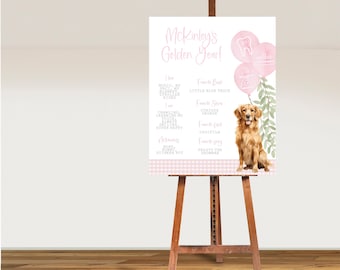 Op de afbeelding: Een roze en wit bord met de tekst "Mckinley's Golden Year!" en een lijst met dingen die het kind leuk vindt, waaronder muziek, dekens en een kleine blauwe vrachtwagen. Het bord bevat ook een aquarel-illustratie van een golden retriever-puppy.