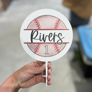 May include: A round, white baseball-themed sign with the name "Rivers" in black script. The sign features a baseball design with red stitching and the number "1". It is held by a white handle.