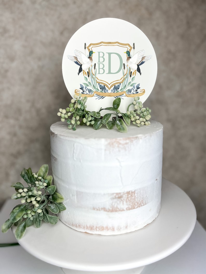 K&ouml;nnte beinhalten: Ein wei&szlig;er Kuchen mit gr&uuml;nem Laub obenauf. Ein runder Kuchentopper mit einem goldenen Schild und den Buchstaben "BD" in Gr&uuml;n befindet sich oben auf dem Laub. Zwei Enten befinden sich auf beiden Seiten des Schildes.